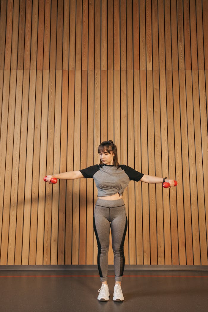 A woman in activewear performs exercises with dumbbells indoors against a wooden wall.