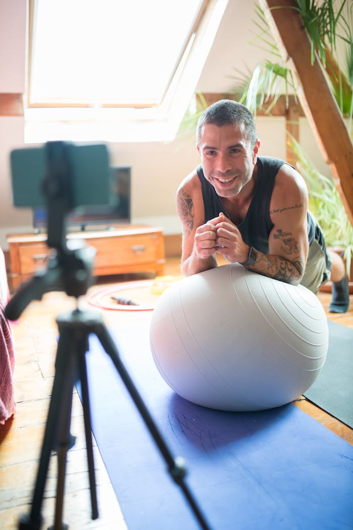 Smiling man exercises on a yoga mat with an exercise ball, capturing workout on video indoors.